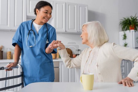 glückliche multirassische Krankenschwester in blauer Uniform, die Hand einer Seniorin mit grauen Haaren hält, während sie neben einem Rollator steht 