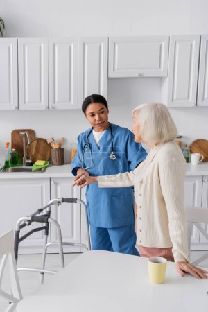 brunette multiracial caretaker in blue uniform supporting senior woman with grey hair while standing near walker 