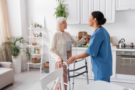 brünette multirassische Krankenschwester in blauer Uniform unterstützt fröhliche Seniorin mit grauen Haaren, die neben Rollator steht 