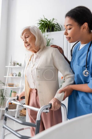 caring multiracial nurse in blue uniform supporting senior woman with grey hair walking with help of walker at home 