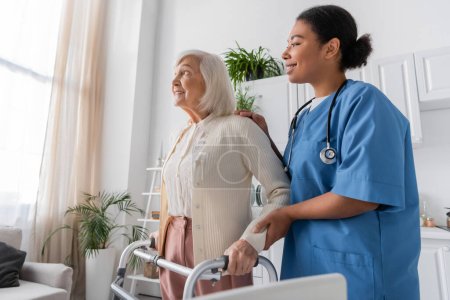 fröhliche multiethnische Krankenschwester in blauer Uniform unterstützt fröhliche Seniorin mit grauen Haaren beim Gehen mit Hilfe eines Rollators zu Hause 