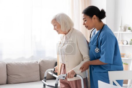 brunette multiracial nurse in uniform supporting retired woman with grey hair walking with walker at home 