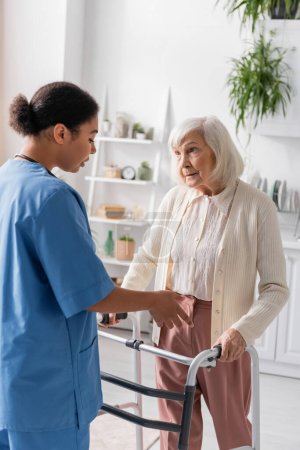 senior woman with grey hair walking with help of walker and looking at brunette multiracial nurse 