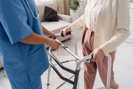 Téléchargez les photos : Vue partielle de l'infirmière multiraciale en uniforme bleu soutenant la femme âgée marchant avec l'aide d'un marcheur à la maison - en image libre de droit