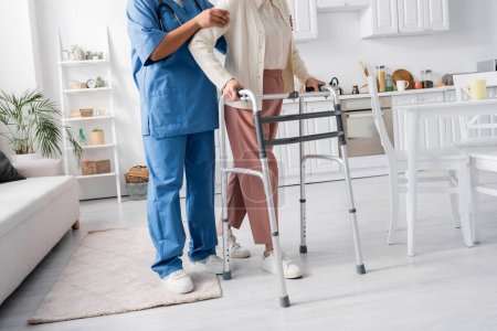 cropped view of multiracial caregiver in uniform supporting senior woman walking with walker in modern apartment 