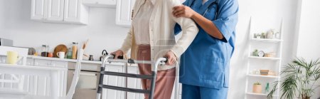 cropped view of multiracial caregiver in uniform supporting senior woman walking with walker in modern apartment, banner 
