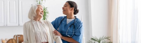happy multiracial nurse in uniform laughing with retired woman at home, banner 