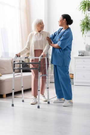 Téléchargez les photos : Pleine longueur de la femme retraitée avec les cheveux gris marchant avec le cadre de marche et regardant l'infirmière multiraciale en uniforme - en image libre de droit