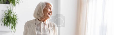 happy senior woman with grey hair looking away while smiling at home, banner 