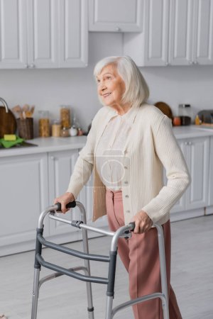 mujer mayor positiva con pelo gris caminando con la ayuda de caminante en casa 