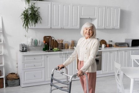 mujer mayor positiva con pelo gris caminando con ayuda de andador en apartamento moderno 