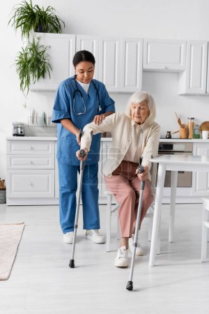 Foto de Longitud completa de la enfermera multirracial en uniforme ayudar a las mujeres mayores que usan muletas para ponerse de pie - Imagen libre de derechos