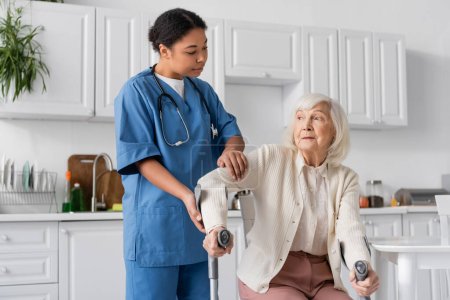 Téléchargez les photos : Infirmière multiraciale en uniforme aidant la femme âgée à utiliser des béquilles pour se lever dans un appartement moderne - en image libre de droit