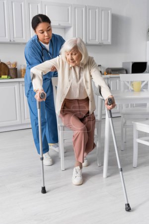 full length of multiracial caregiver in uniform helping senior woman using crutches to stand up at home 