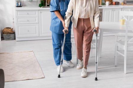 Téléchargez les photos : Vue recadrée de la femme âgée utilisant des béquilles tout en marchant près de l'infirmière à la maison - en image libre de droit