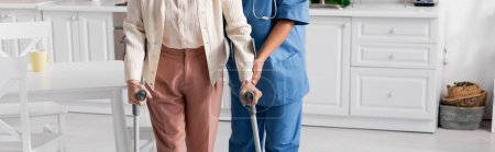 cropped view of senior woman using crutches while walking near nurse at home, banner 