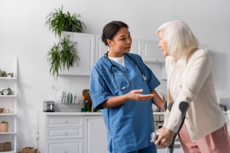 Rentnerin mit grauen Haaren auf Krücken und im Gespräch mit Krankenschwester zu Hause 