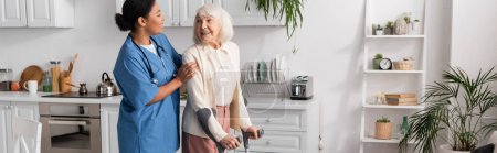 Téléchargez les photos : Femme âgée gaie avec les cheveux gris à l'aide de béquilles et de marcher près infirmière multiraciale à la maison, bannière - en image libre de droit