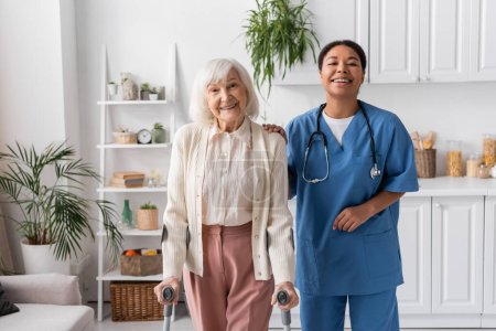 positive Rentnerin mit grauen Haaren auf Krücken beim Gehen in der Nähe einer glücklichen multirassischen Krankenschwester zu Hause 