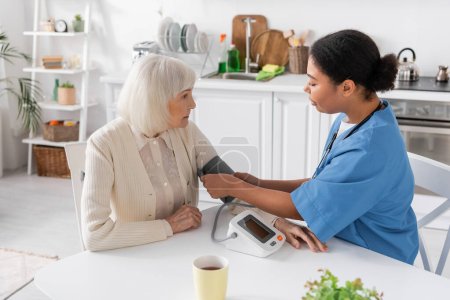multiracial nurse measuring blood pressure of senior woman with grey hair 