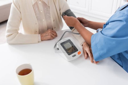 Téléchargez les photos : Vue recadrée de l'infirmière multiraciale mesurant la pression artérielle de la femme âgée - en image libre de droit