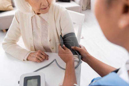 partial view of multiracial nurse measuring blood pressure of senior woman with grey hair 