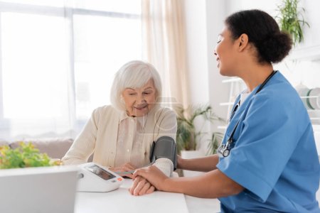 joyful multiracial nurse measuring blood pressure of senior woman with grey hair 