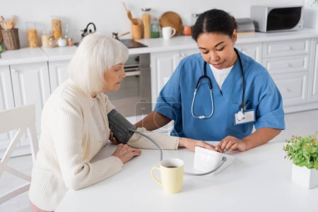 enfermera multirracial que mide la presión arterial de la mujer mayor con el pelo gris al lado de la taza de té en la mesa 