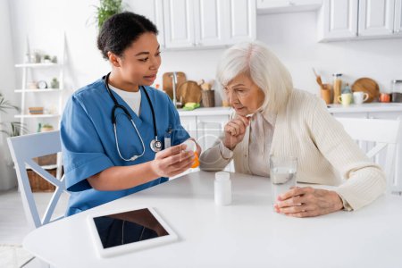 Krankenschwester zeigt Seniorin neben digitalem Tablet Medikament auf Tisch 