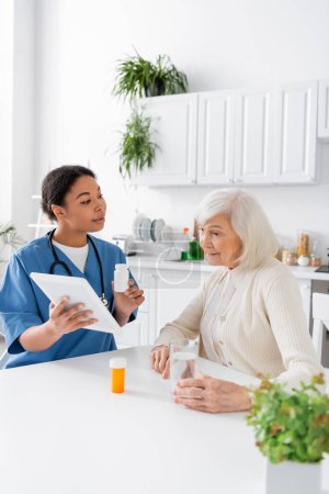 brünette multirassische Krankenschwester mit digitalem Tablet und Medikamenten in der Hand, während sie mit einer Rentnerin mit grauen Haaren spricht