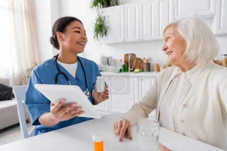 glückliche multiethnische Krankenschwester mit digitalem Tablet und Medikamenten in der Hand, während sie mit einer Rentnerin mit grauen Haaren spricht