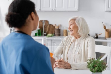 mujer jubilada feliz con el pelo gris mirando lejos cerca de enfermera multiracial en primer plano borrosa 