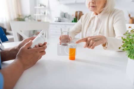 Ausgeschnittene Ansicht einer Seniorin, die in der Nähe einer Krankenschwester auf eine Flasche mit Medikamenten zeigt, im verschwommenen Vordergrund 