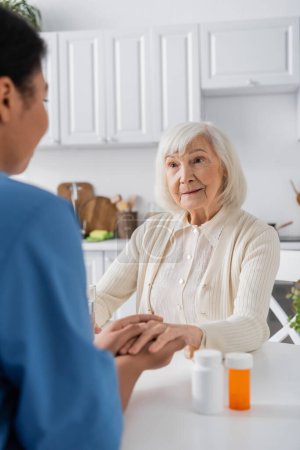 Foto de Enfermera multirracial borrosa cogida de la mano mientras reconforta a la mujer mayor junto a la medicación en la mesa - Imagen libre de derechos
