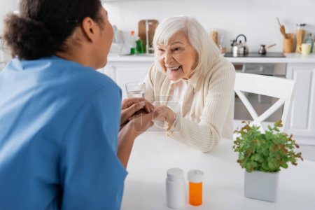 Krankenschwester hält Hand in Hand mit glücklicher Seniorin neben Medikamenten auf Tisch 
