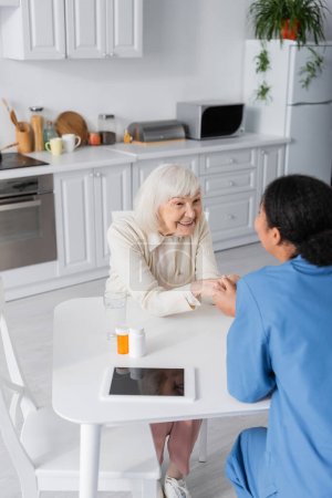 multiracial nurse holding hands with cheerful senior woman next to medication on table 