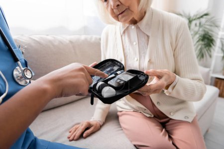 Téléchargez les photos : Vue recadrée de l'infirmière multiraciale en uniforme pointant vers le kit diabète près de la femme âgée assise sur le canapé - en image libre de droit
