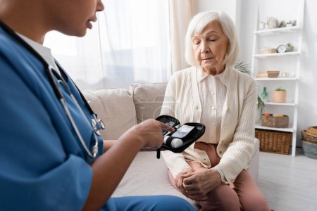 Pflegerin in Uniform zeigt auf Diabetes-Set neben Seniorin, die auf Sofa sitzt 