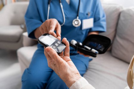 Teilansicht einer Krankenschwester, die auf Glukometer in der Hand einer Seniorin zeigt