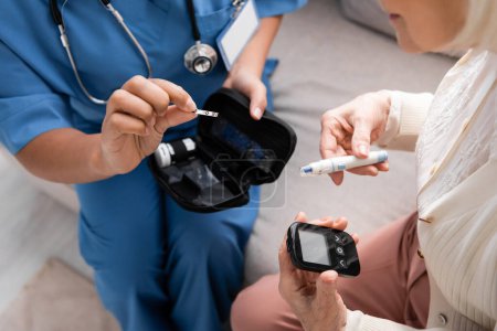zugeschnittene Ansicht einer multirassischen Krankenschwester in Uniform mit Teststreifen in der Nähe einer älteren Frau mit Lanzetstift und Glukometer 