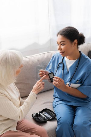 brünett multiracial krankenschwester in uniform holding teststreifen und glucometer in der nähe von senior woman 