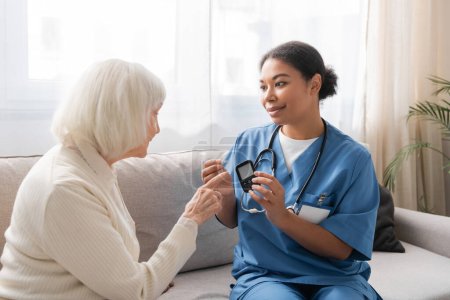 glücklich multirassische Krankenschwester in Uniform hält Teststreifen und Glukometer in der Nähe von Seniorin 