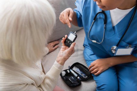 cropped view of multiracial caregiver in uniform pointing at glucometer near senior woman with grey hair 