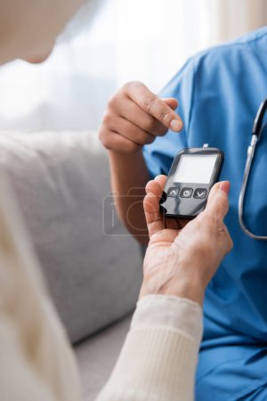 cropped view of multiracial nurse pointing at glucometer with test strip near senior woman