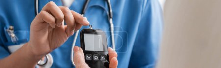 cropped view of multiracial nurse pointing with finger at glucometer with test strip near senior woman, banner 