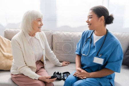 Téléchargez les photos : Heureuse femme âgée tenant glucomètre près infirmière multiraciale en uniforme bleu - en image libre de droit