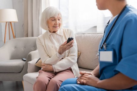 Glückliche Seniorin blickt in blauer Uniform auf Glukometer in der Nähe einer Krankenschwester 