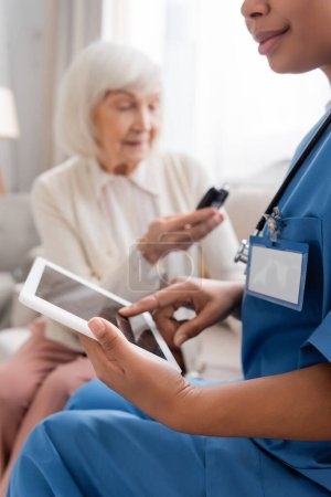 Téléchargez les photos : Infirmière multiraciale utilisant tablette numérique avec écran blanc près de la femme âgée sur fond flou - en image libre de droit