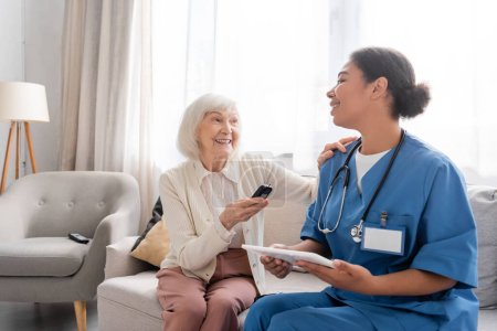 Foto de Alegre anciana con pelo gris sosteniendo glucosímetro cerca feliz enfermera multirracial utilizando tableta digital - Imagen libre de derechos