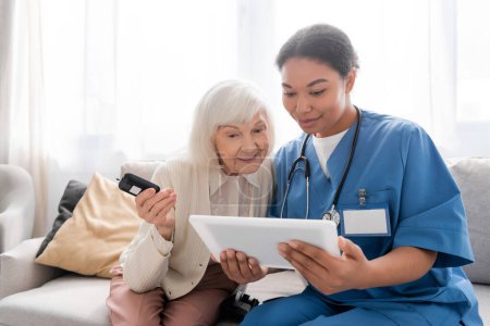 Foto de Alegre anciana con pelo gris sosteniendo el glucosímetro y mirando la tableta digital cerca de feliz enfermera multirracial - Imagen libre de derechos